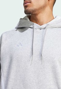 Lys grå hettegenser laget av myk stoff, med snorhette, hvite detaljer og en hvit Adidas-logo på brystet.