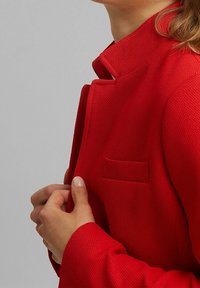 Rode textuurige jas met een opstaande kraag en een klein zakje aan de linkerzijde. Het materiaal lijkt geweven en toont een levendige kleur.