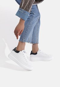 Sneakers bianchi con un accento nero sul tallone, texture liscia, punta arrotondata e suola rialzata, abbinate a jeans cropped blu chiaro.