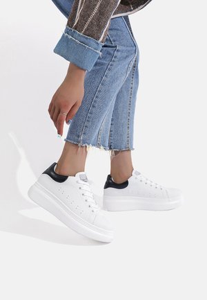 Sneakers bianchi con un accento nero sul tallone, texture liscia, punta arrotondata e suola rialzata, abbinate a jeans cropped blu chiaro.