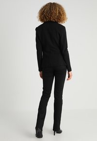 Žena s kudrnatými vlasy, která má na sobě přiléhavý černý blazer, černé kalhoty a černé lodičky, stojí zády k objektivu na jednobarevném pozadí.