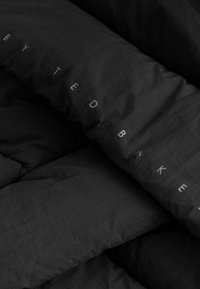 Schwarze gesteppte Bettwäsche mit glattem Textur, auf einer Seite mit dem Text "by TED BAKER" in dezent grauer Schrift.