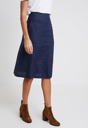 Femme portant une jupe bleu marine longueur genou, un haut blanc et des bottines en daim marron, debout sur un fond uni.