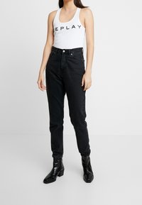 Jeans neri a vita alta con una vestibilità affusolata, caratterizzati da un lavaggio scuro, tasche frontali e chiusura a bottone. Abbinati a un top bianco senza maniche.