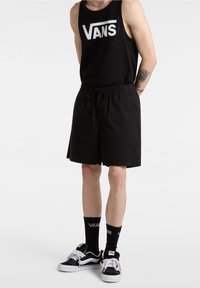 Person som bär en svart linne med "VANS"-logotyp, svarta shorts och svarta strumpor med skor. Bakgrunden är helt vit.