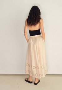 Gonna beige, lunga fino al ginocchio con vita arricciata, caratterizzata da motivi a taglio circolare lungo l'orlo. Indossata con un crop top nero e sandali.