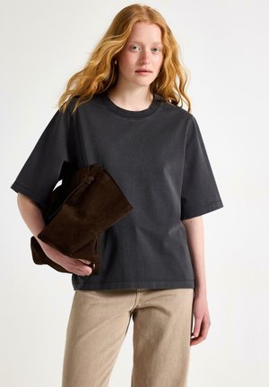 Ung kvinde med langt rødt hår iført en løs mørkegrå T-shirt og beige bukser, der holder en foldet mørkebrun ruskindsstøvle.