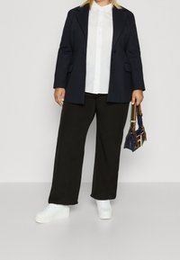 Blazer bleu marine foncé, chemise blanche à col, pantalon noir large et baskets blanches. Petit sac avec une anse multicolore à la main.