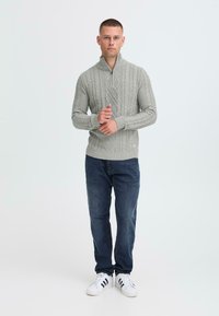 Light gray knitted sweater with a high collar and cable knit design, paired with dark blue jeans and white sneakers with black stripes.