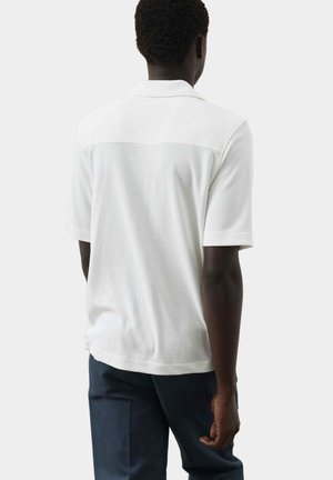 Mann trägt weißes, strukturiertes Poloshirt mit kurzen Ärmeln und dunkelblaue Hose, steht mit dem Rücken zur Kamera vor einfarbigem Hintergrund.