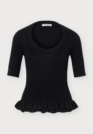 Femme aux longs cheveux blonds portant un haut noir à manches courtes avec un ourlet à volants, une longue jupe bordeaux fendue sur le côté et des bottes noires, debout bien droite.
