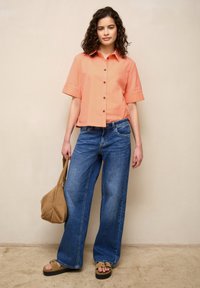 Femme debout aux cheveux bouclés, portant une chemise pêche à manches courtes boutonnée, un jean bleu à jambes larges, des sandales beige et tenant un sac beige.
