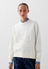 Weißer Pullover mit einem geprägten Design von Totenkopf und gekreuzten Knochen. Texturiertem Stoff mit gerippten Bündchen und Saum, übereinander getragen über einem blauen Shirt.