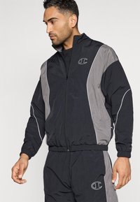 Mann in einer schwarzen und grauen Kapuzenjacke und Jogginghose mit weißer Paspelierung und Champion-Logo auf der Brust und dem Bein, der nach links schaut.