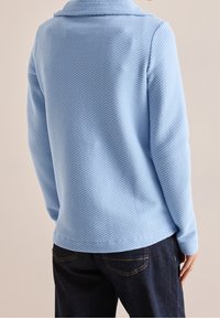 Persona con una camiseta de manga larga texturizada de color azul claro con cuello y jeans de mezclilla oscuros, de pie contra un fondo beige liso.