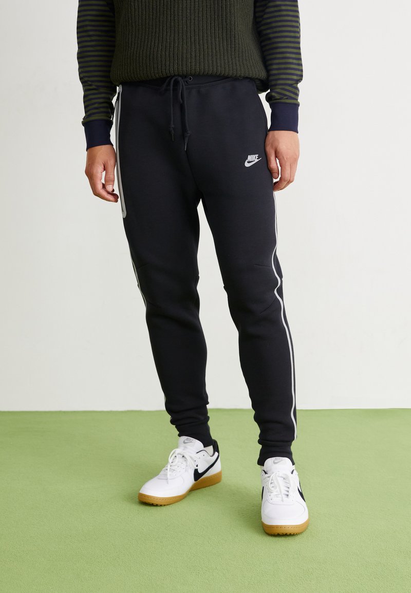 Osoba u crnim Nike trenirkama s bijelim bočnim prugama i bijelim Nike tenisicama stoji na svijetlozelenom podu.