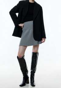 Personne portant un blazer noir, un top noir, une jupe en gris à côtes et des bottes en cuir noir jusqu'aux genoux, se tenant avec la main sur la hanche.