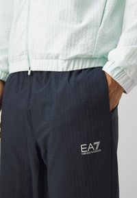 Casaco xadrez verde claro com punhos elásticos, combinado com calções azul escuro com um padrão xadrez subtil e logo EA7.