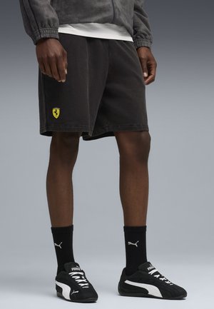 Sorte bomulds shorts med et gult Ferrari-logo, båret med sorte Puma-sko og sokker. Teksturen ser blød ud med en afslappet pasform.