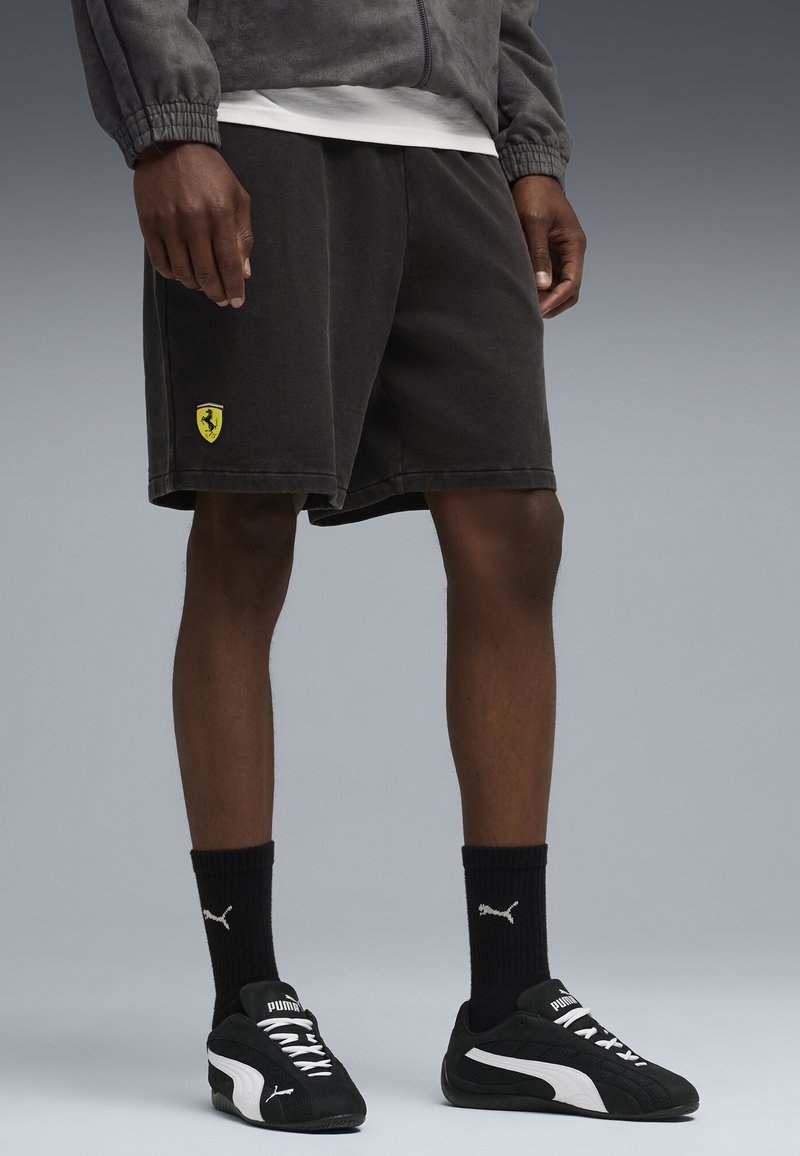 Sorte bomulds shorts med et gult Ferrari-logo, båret med sorte Puma-sko og sokker. Teksturen ser blød ud med en afslappet pasform.