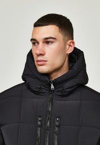 Schwarze gesteppte Daunenjacke mit hohem Kragen und durchgehendem Reißverschluss, ausgestattet mit Kordelzugverstellungen und schwarzen Hardware-Akzenten.