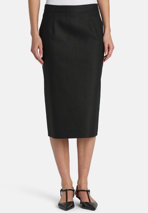 Nő, aki térdig érő fekete ceruzaszoknyát és fekete, hegyes orrú T-pántos magassarkút visel, kezeit lazán oldalán tartva áll.