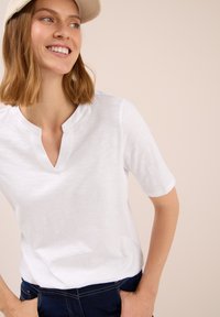 Smilende kvinde med skulderlangt lysebrunt hår iført en hvid kortærmet top og mørkeblå jeans, hænderne i lommerne, ser til venstre.
