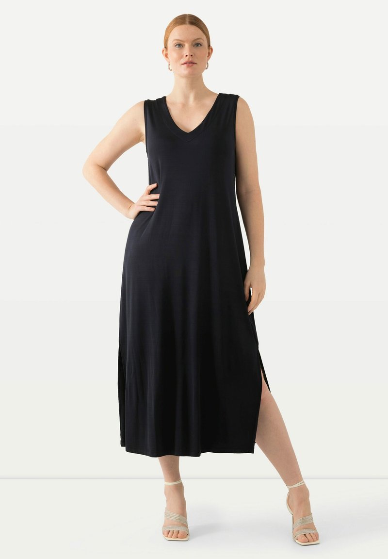 Abito lungo senza maniche di colore nero con scollatura a V, spacchi laterali e una superficie liscia. Abbinato a sandali beige con cinturini.