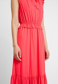 Korallenrosa Kleid mit gerafftem Taillenbereich, rüschierten Akzenten an den Ärmeln und am Saum, aus weichem Stoff gefertigt, mit einem fließenden Design.