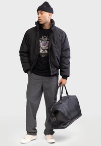 Zwarte pufferjack, zwart graphic t-shirt, grijze broek en off-white sneakers. Houdt een grote zwarte leren sporttas vast.