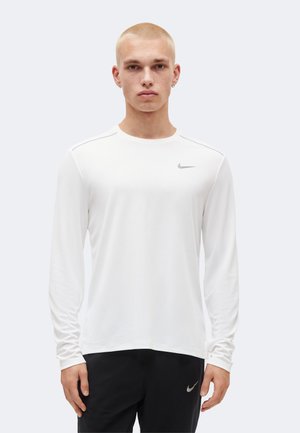 Ung mand iført en hvid langærmet Nike-trøje og sorte Nike-bukser, stående og vendt fremad mod en ensfarvet baggrund.