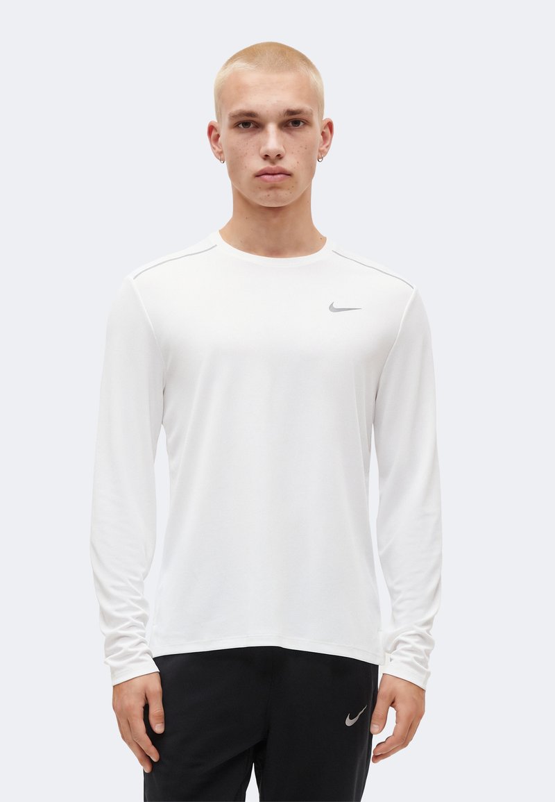 Νέος άνδρας που φοράει λευκό μακρυμάνικο μπλουζάκι Nike και μαύρο παντελόνι Nike, στέκεται και κοιτάζει μπροστά σε απλό φόντο.