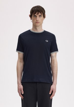 Uomo con capelli corti castani che indossa una maglietta nera con bordi bianchi e un piccolo logo con corona d'alloro sul petto, in piedi davanti a uno sfondo semplice.