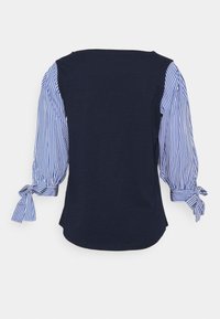 Marinblus med blå och vita randiga trekvart ärmar, samlade ärmslut knutna med rosetter, visad bakifrån mot en vit bakgrund.