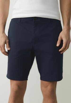 Mężczyzna w granatowych, dopasowanych szortach i białej koszulce bez rękawów, stojący z rękami luźno opuszczonymi wzdłuż ciała na jednolitym tle.