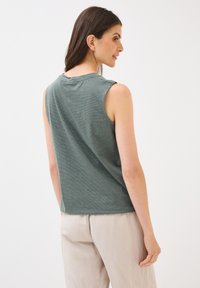 Modèle de dos, portant un haut sans manches à rayures vertes et un pantalon beige clair, cheveux lâchés, avec un fond neutre.