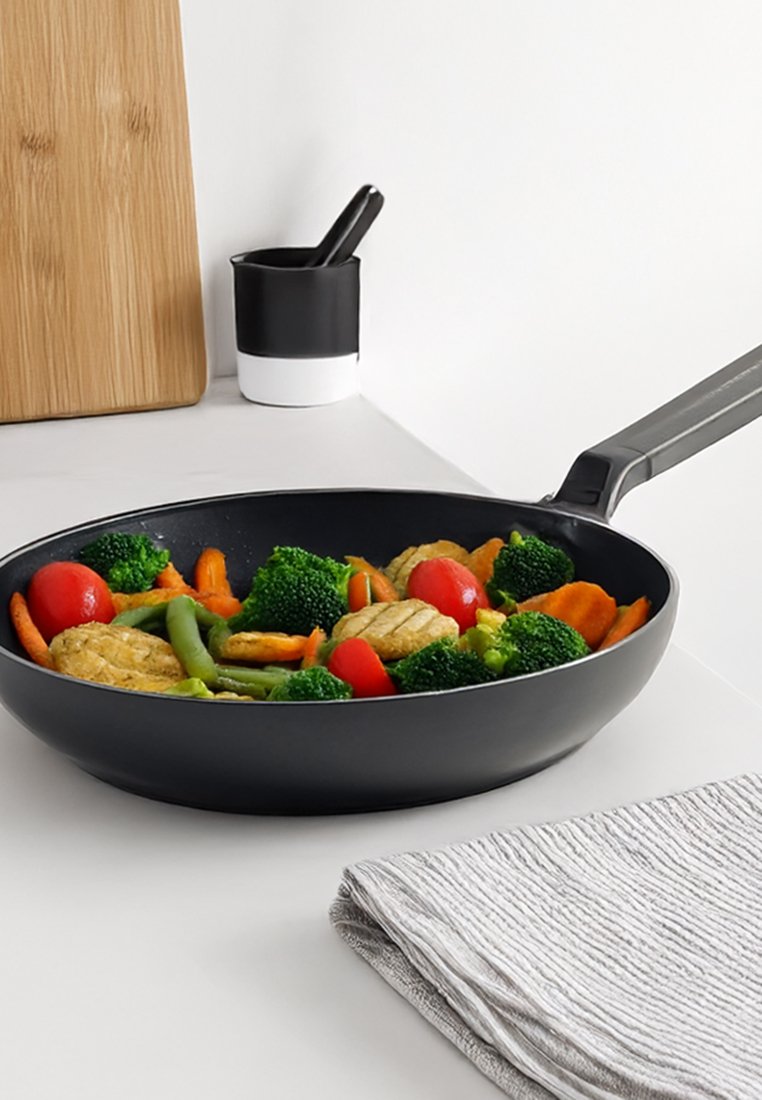 Schwarze antihaftbeschichtete Bratpfanne mit gemischtem Gemüse: Brokkoli, Karotten, grüne Bohnen und Kirschtomaten. Leicht graues, strukturiertes Geschirrtuch in der Nähe.