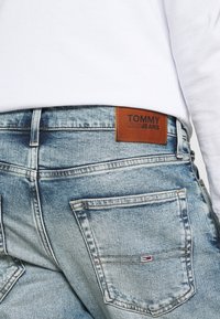 Jean en denim bleu clair avec une finition légèrement délavée, comportant une étiquette en cuir marron portant la mention "TOMMY JEANS" sur la ceinture.