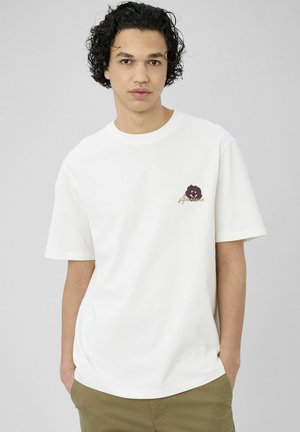 Jeune homme aux cheveux noirs bouclés portant un t-shirt blanc avec une petite fleur sombre et un texte sur la poitrine, ainsi qu'un pantalon vert olive.