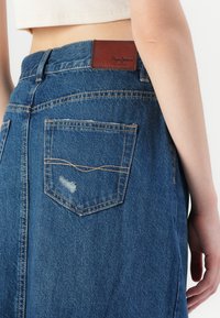 Blue denim skirt featuring a high waist, brown leather logo patch, and a back pocket with a distressed detail and contrasting stitching.