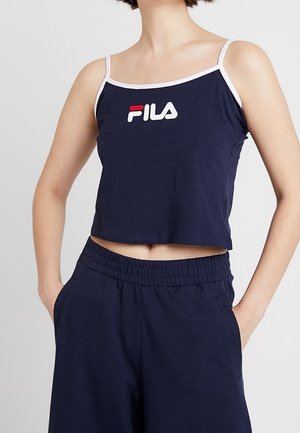 Woman wearing a navy blue FILA crop top with white trim and matching navy blue elastic waist pants, hands in pockets, against white background.