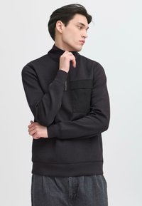 Tricou negru cu guler înalt, buzunar frontal cu fermoar, manșete și tiv din jersey. Asortat cu pantaloni gri, largi, dintr-un material texturat.