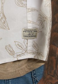 Short-sleeved shirt featuring a light beige fabric with subtle floral pattern, and a woven brand label. The hem has a contrasting beige texture.
