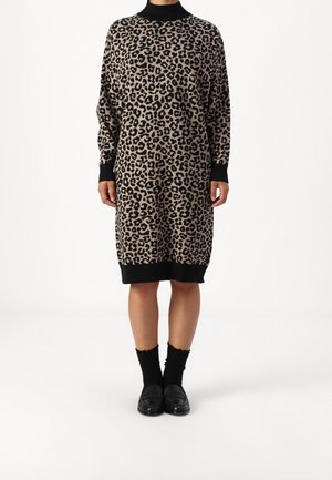 Person trägt ein knielanges Pulloverkleid mit Leopardenmuster, schwarzen Bündchen, Kragen und Saum, schwarze Socken und schwarze Loafers und steht vor einem weißen Hintergrund.