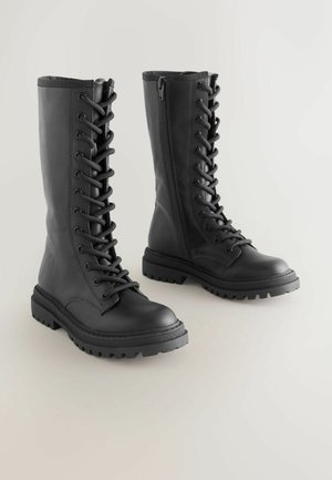 Crne kožne čizme s vezicama i visokim okovratnikom, bočnim patentnim zatvaračem, robusnom potplatom i teksturiranom površinom. Imaju okrugli vrh i izdržljive šavove.