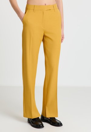 Personne portant un pantalon ajusté jaune moutarde avec des poches latérales et des mocassins en cuir noir, debout sur un fond blanc uni.