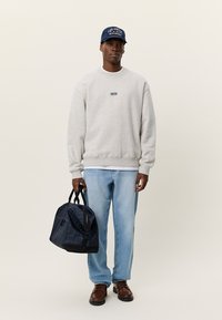 Άνδρας που φορά ανοιχτό γκρι φούτερ, μπλε καπέλο, ανοιχτό μπλε τζιν, καφέ παπούτσια, κρατώντας σκούρο μπλε καρό σακ βουαγιάζ πάνω σε μπεζ φόντο.