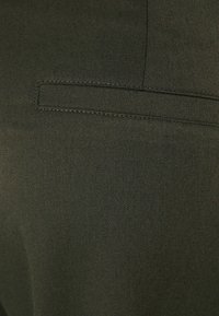 Olivgrön tyg med en fin twillstruktur och en enkel kanteficka. Slät yta med subtila sömmar som skapar strukturella linjer.