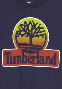 Marinblå bomullströja med ett stort cirkulärt mönster i gult och orange, med en svart träsilhuett och "Timberland"-logotypen i rött nedan.