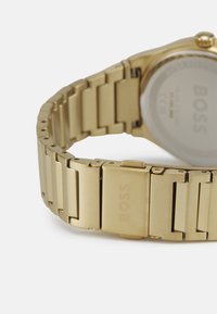 Reloj de metal dorado con un diseño de pulsera enlazada. Cuenta con un cierrem rectangular y el nombre de la marca grabado, textura suave y acabado pulido.